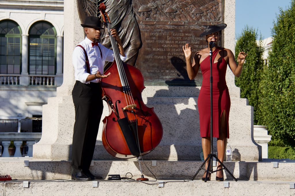 acute inflections, woman in red dress and hat singing with man in button down shirt playing cello