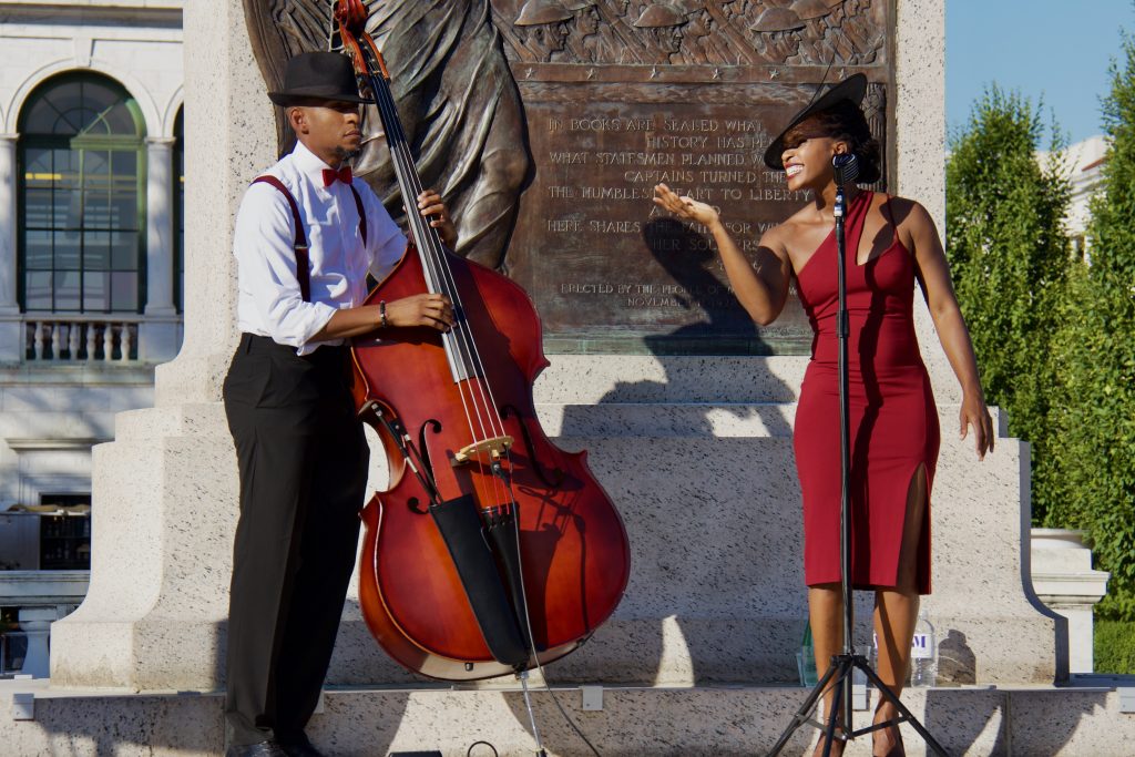 acute inflections, woman in red dress and hat singing with man in button down shirt playing cello