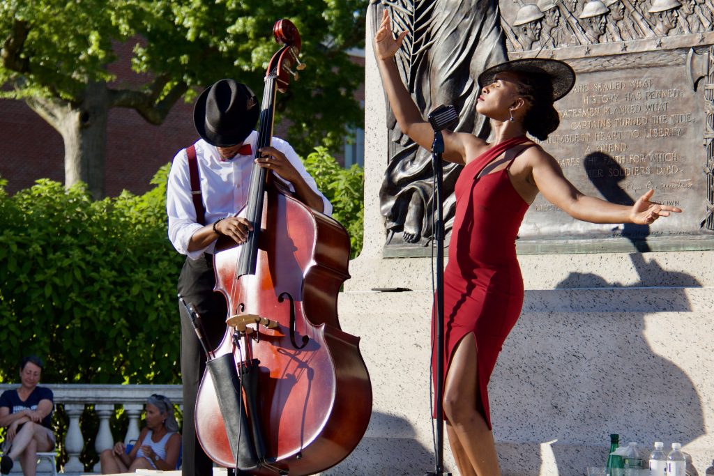 acute inflections, woman in red dress and hat singing with man in button down shirt playing cello