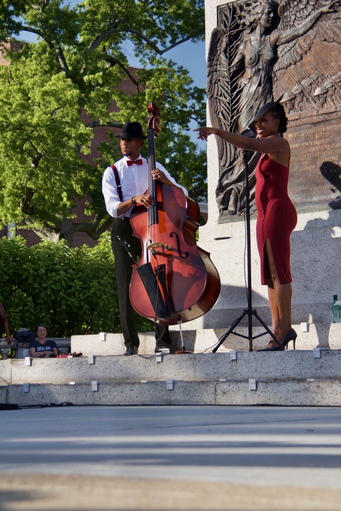 acute inflections, woman in red dress and hat singing with man in button down shirt playing cello